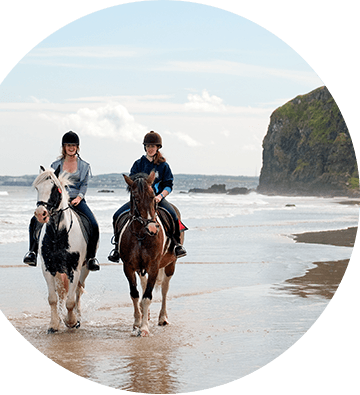 Two women riding horseback on the shores with cliffs in the background.